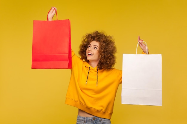 Retrato de feliz hipster de cabelos cacheados feliz com capuz levantando sacolas de compras com área em branco, copie o espaço em pacotes para texto publicitário. tiro de estúdio interno, comercial de loja de moda