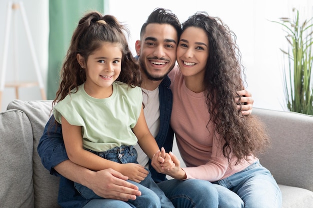 Retrato de feliz jovem família de três do Oriente Médio com filha relaxando juntos em casa, mãe, pai e filho olhando e sorrindo para a câmera enquanto abraçando no sofá na sala de estar