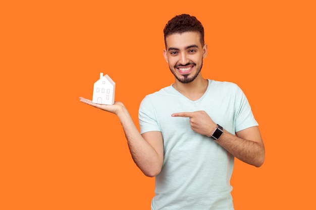 Retrato de feliz jovem morena com barba em t-shirt branca em pé apontando para a casa de papel na palma da mão e sorrindo para a câmera, compra de casa. tiro de estúdio interno isolado em fundo laranja