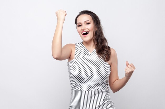 Retrato de feliz linda jovem morena com maquiagem e vestido listrado em pé e olhando para a câmera com os punhos e comemorando sua vitória. tiro de estúdio interno, isolado em fundo cinza.