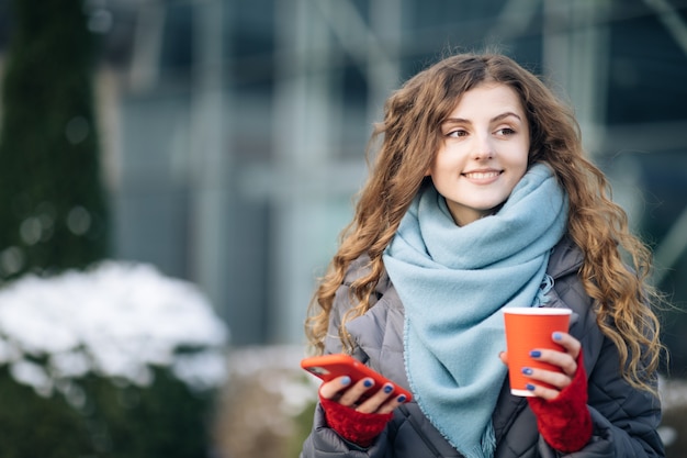 Retrato de feliz linda mulher encaracolada com xícara de café para viagem usar mensagens de texto do telefone móvel.