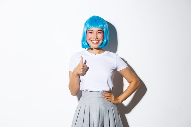 Retrato de feliz menina asiática na peruca azul, mostrando o polegar para cima em aprovação, recomendo algo, de pé.