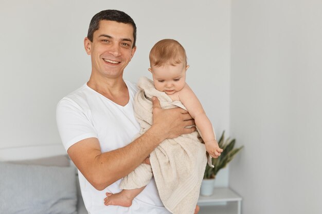 Retrato de feliz pai adulto jovem positivo com sua filha criança brincando juntos em casa, criança sendo enrolada em uma toalha depois do banheiro, homem olhando sorrindo para a câmera.