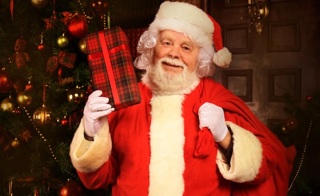 Retrato de feliz papai noel sentado em seu quarto em casa perto da árvore de natal com caixa de presente.