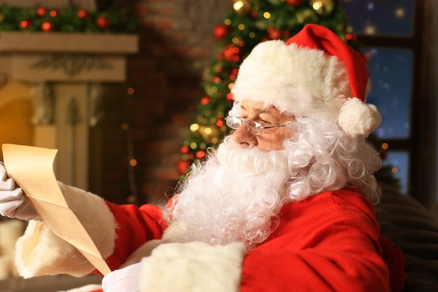 Retrato de feliz papai noel sentado em seu quarto em casa perto da árvore de natal e lendo a carta de natal ou a lista de desejos.