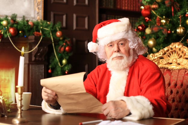 Retrato de feliz Papai Noel sentado em seu quarto em casa perto da árvore de Natal e lendo a carta de Natal ou a lista de desejos.