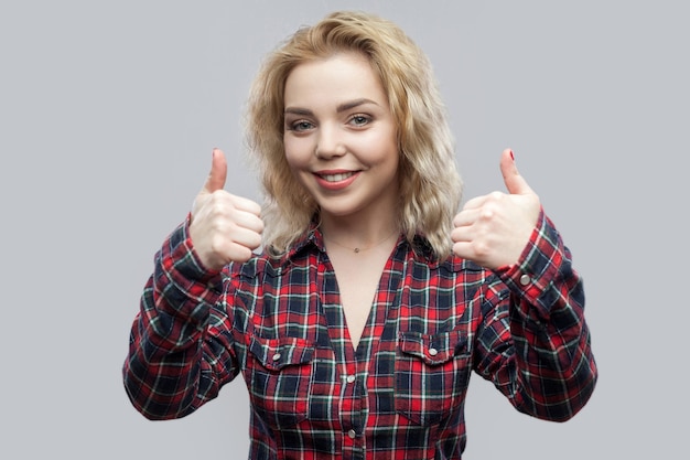 Retrato de feliz satisfeito linda loira jovem em casual camisa quadriculada vermelha em pé e olhando para a câmera com um sorriso largo e polegares para cima. tiro do estúdio, isolado no fundo cinza.