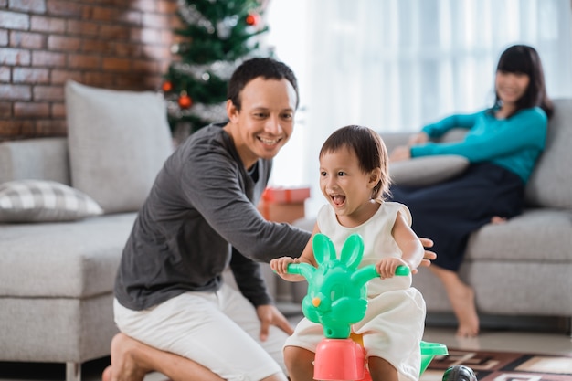 Retrato de filha brincando de triciclo com o pai ajudado e a mãe sentada em um sofá atrás