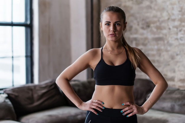 Retrato de forte desportivo magro desportivo caucasiano jovem vestindo sutiã esportivo preto em pé com as mãos nos quadris, olhando para a câmera no estúdio loft