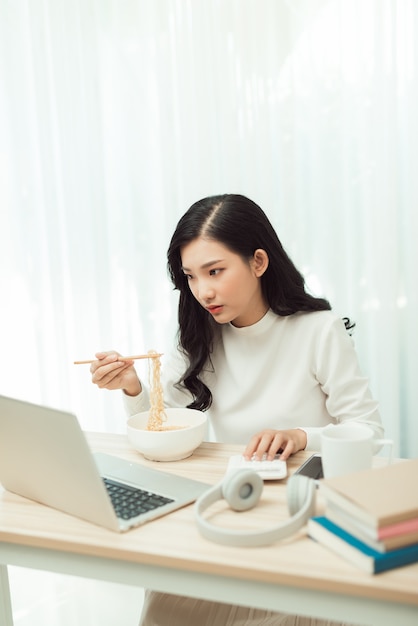 Retrato de freelancer jovem asiática, trabalhando com o laptop em sua casa e comendo comida asiática com comida para viagem.