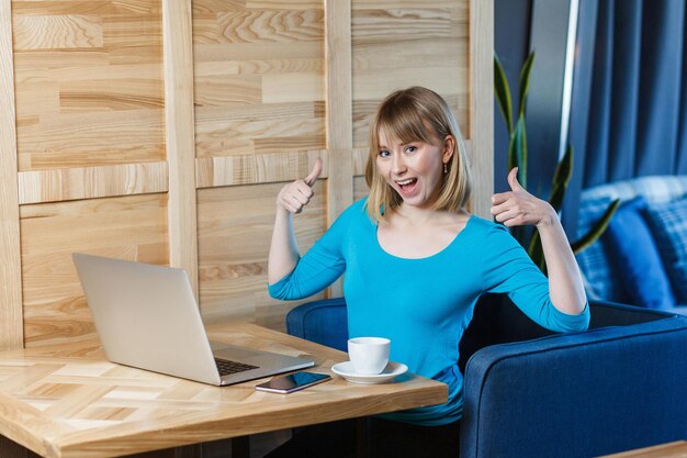 Foto retrato de freelancer jovem positivo atraente com cabelo loiro bob na blusa azul estão sentados no café e trabalhando no laptop com sorriso e aparecendo o polegar, olhando para a câmera.