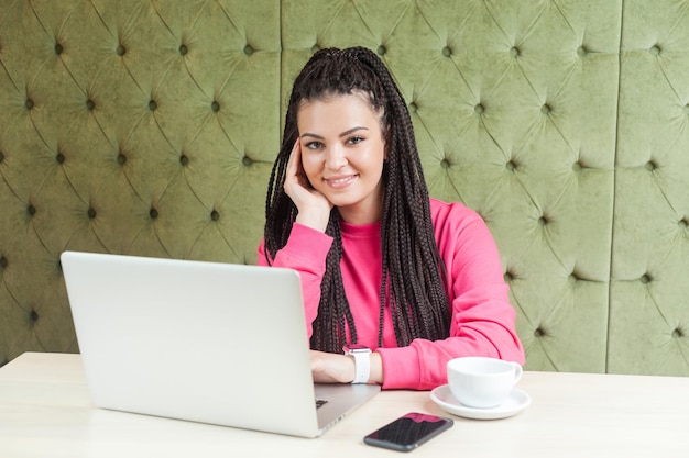 Retrato de freelancer linda jovem satisfeita com penteado de dreadlocks preto na blusa rosa, sentado no café, trabalhando no laptop com sorriso, segurando uma mão no queixo, olhando para a câmera.