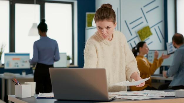 Retrato de funcionário de inicialização focado olhando para laptop e gráficos comparando estatísticas de vendas em pé na mesa. Mulher no escritório ocupado classificando documentos comerciais, analisando a análise de dados.