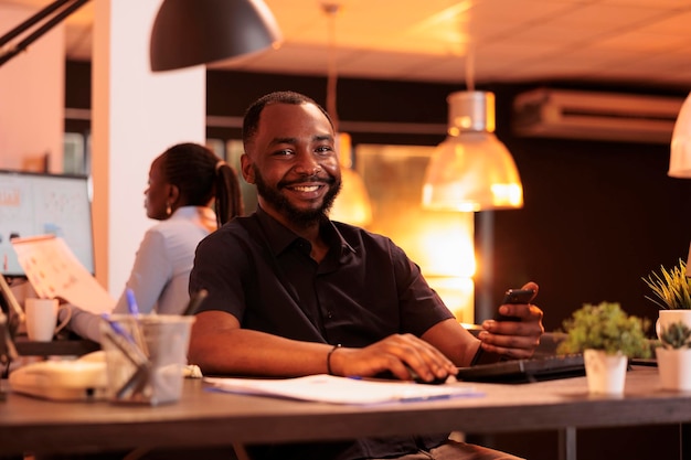 Retrato de funcionário do sexo masculino usando aplicativo de smartphone no trabalho de escritório, trabalhando com telefone celular e computador para criar relatório de negócios. Navegando rede de mídia social para fazer pesquisa de papelada.