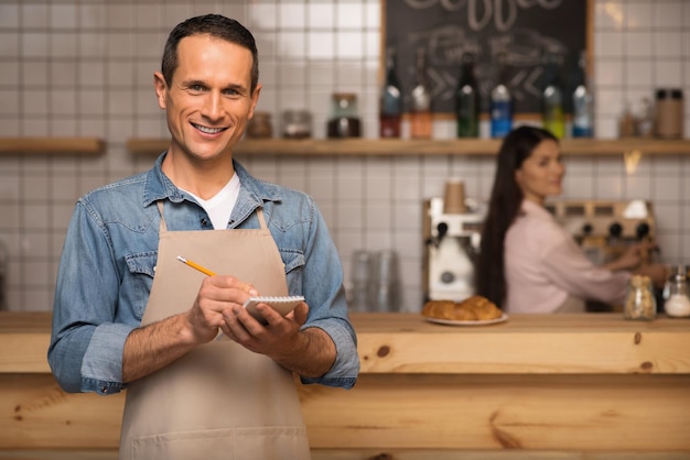 Retrato de garçom sorridente tomando notas e olhando para a câmera com garçonete no fundo