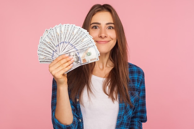 Retrato de garota de sorte feliz na camisa quadriculada segurando muito dinheiro e sorrindo para a câmera mostrando notas de dólar na mão desfrutando de sucesso financeiro tiro de estúdio interior isolado no fundo rosa