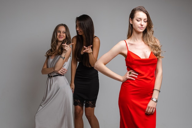 retrato de garotas fofoqueiras em vestidos prateados e pretos zombando de uma linda garota infeliz com longos cabelos loiros em um vestido vermelho em pé em primeiro plano e olhando para baixo com tristeza