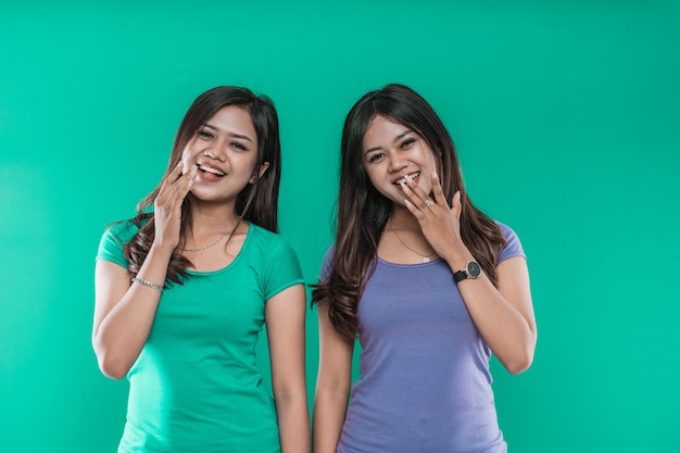 Retrato de gêmeas jovens asiáticas rindo enquanto cobrem a boca com as mãos isoladas sobre fundo verde