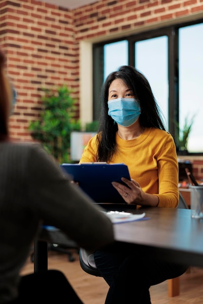 Retrato de gerente asiático usando máscara facial médica para prevenir infecção por coronavírus enquanto entrevistava mulher em potencial para aplicativo de gerente no escritório de inicialização. conceito de negócios