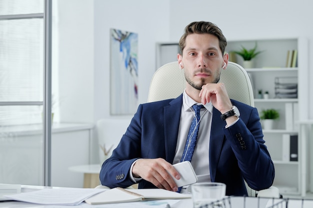 Retrato de gerente de negócios sério em fones de ouvido, sentado na mesa e segurando um telefone moderno no escritório
