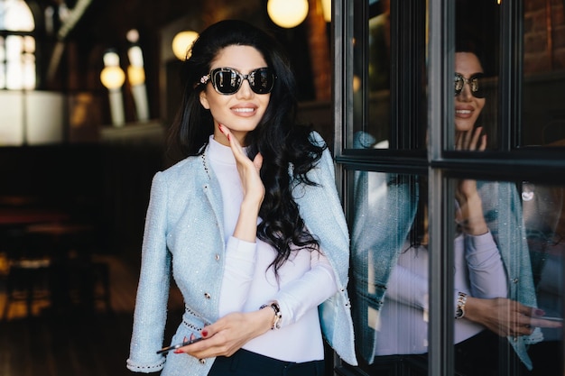 Retrato de glamour jovem com cabelos ondulados escuros usando óculos escuros e blusa branca com jaqueta elegante com manicure vermelha em pé dentro de casa segurando smartphone esperando o namorado no café