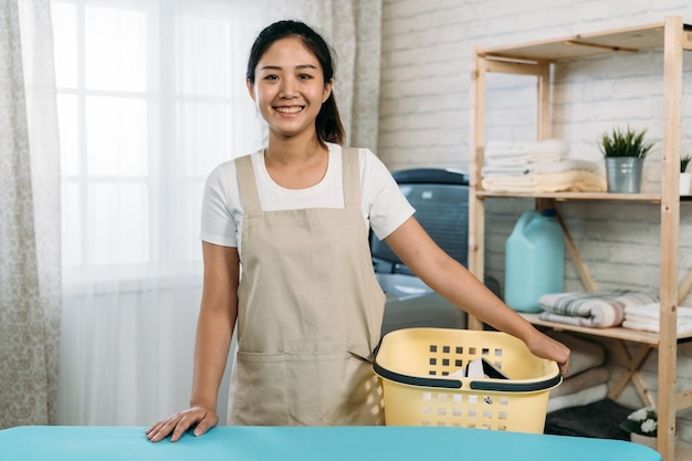 retrato de governanta feminina asiática confiante carregando uma cesta no avental está olhando para a câmera com um sorriso em uma lavanderia brilhante em casa.