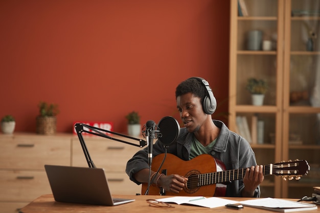 Retrato de grande angular de um homem afro-americano talentoso cantando ao microfone e tocando violão enquanto grava música no estúdio, copie o espaço