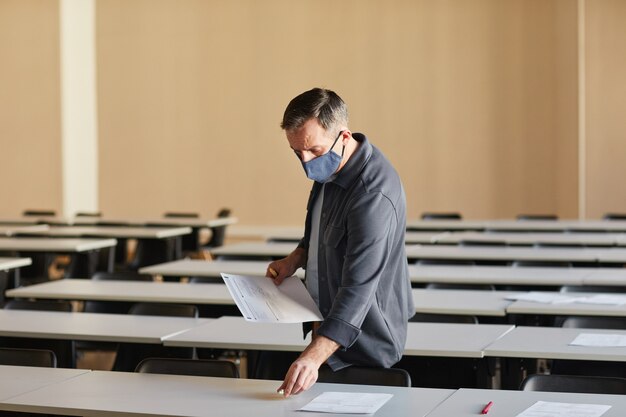 Retrato de grande angular de um professor universitário maduro usando máscara enquanto distribui as provas no auditório da escola iluminado pela luz solar, copie o espaço