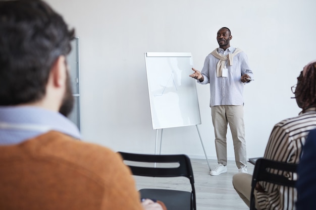 Retrato de grande angular de um treinador de negócios afro-americano conversando com o público em uma conferência ou seminário educacional enquanto está de pé ao lado do quadro branco e gesticulando, copie o espaço