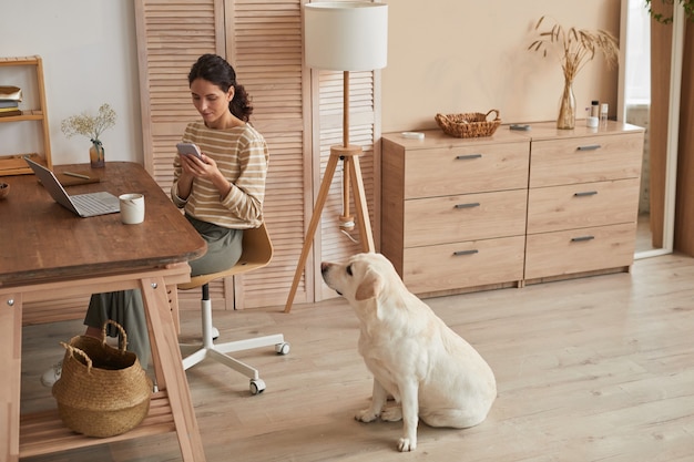 Retrato de grande angular em tons quentes de mulher jovem e moderna usando o smartphone no escritório em casa com um cachorro sentado ao lado dela em um interior aconchegante, copie o espaço