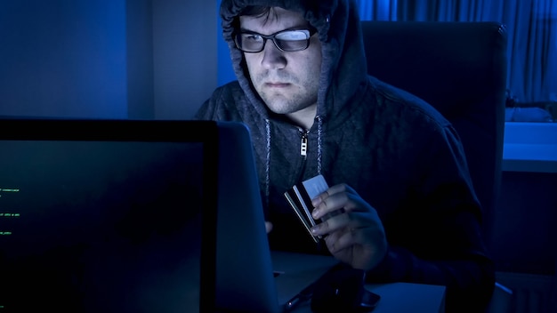 Retrato de hacker segurando cartões de crédito trabalhando no computador à noite