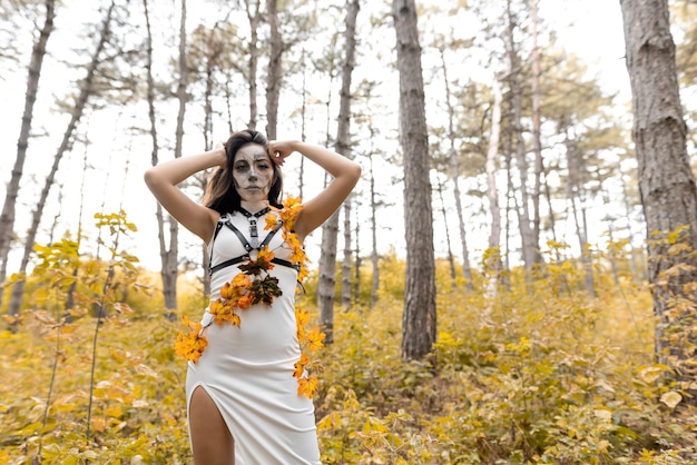 Retrato de Halloween de uma jovem linda em um vestido branco com maquiagem no rosto Posando na floresta de outono Maquiagem de rosto de esqueleto O dia dos mortos
