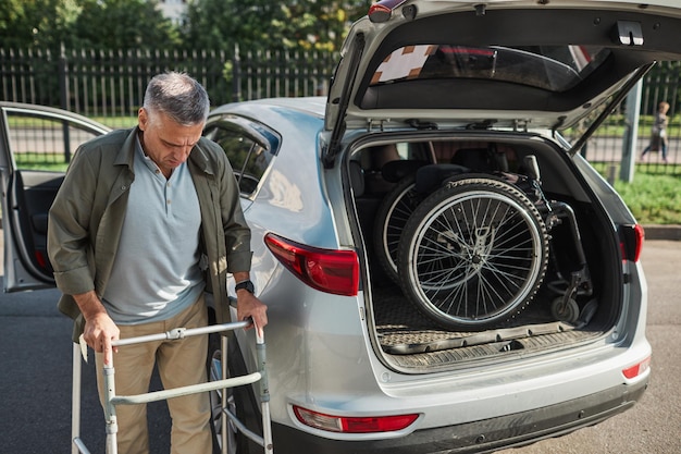 Retrato de homem adulto com deficiência, descarregando cadeira de rodas do porta-malas do carro em um estacionamento ao ar livre ...