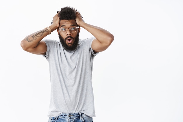 Foto retrato de homem afro-americano chocado e assustado de óculos, segurando o cabelo, boca aberta, dobrando os lábios e erguendo as sobrancelhas em uma expressão facial desesperada, sendo abalado e frustrado, começando a entrar em pânico