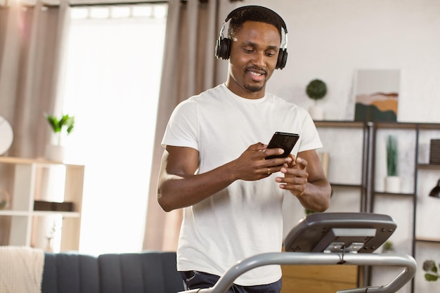 Retrato de homem afro-americano em camiseta e fones de ouvido com telefone na mão na esteira