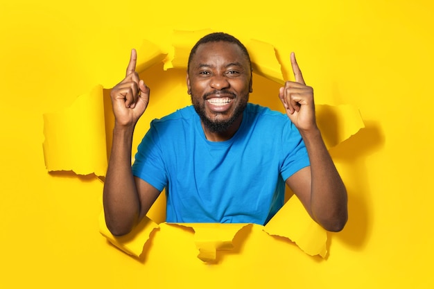 Retrato de homem afro-americano excitado apontando dedos para cima e sorrindo parado em fundo amarelo de buraco de papel rasgado