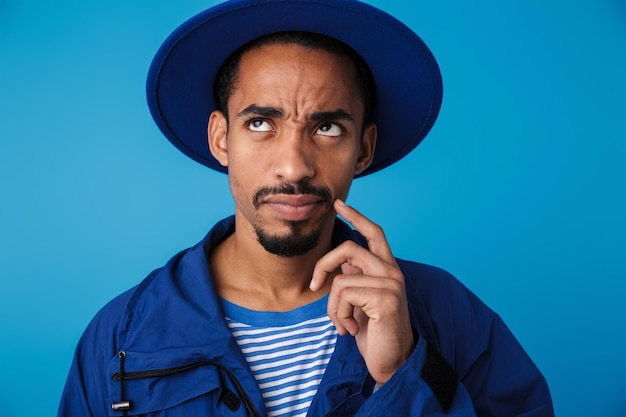 Retrato de homem afro-americano sério usando chapéu, pensando e olhando para cima, isolado no azul