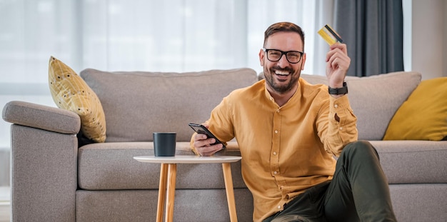 Retrato de homem alegre com cartão de crédito usando telefone celular na sala de estar em casa