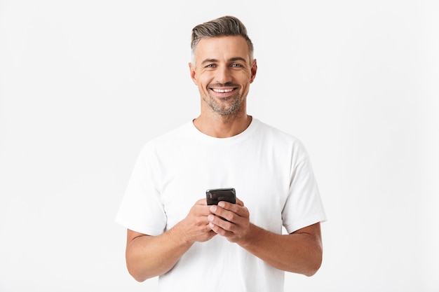 Retrato de homem alegre dos anos 30 vestindo camiseta casual segurando e usando smartphone isolado no branco