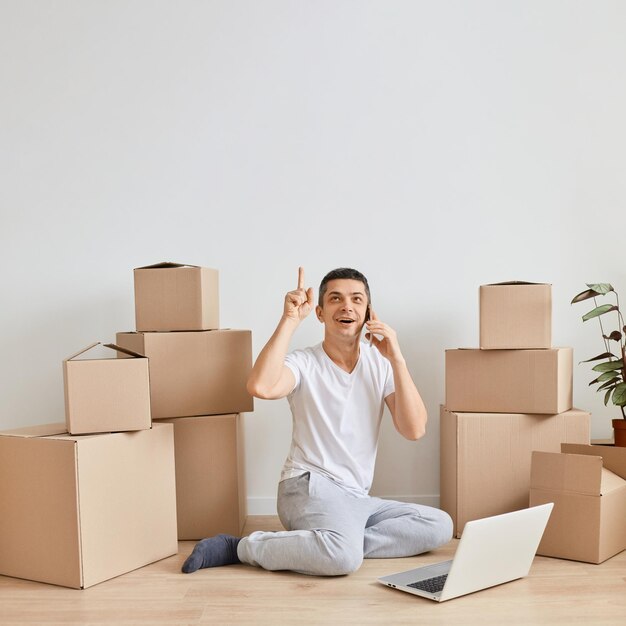 Retrato de homem animado sentado no chão e trabalhando no laptop em um novo apartamento com caixas de papelão desembaladas tendo trabalho online levantou o dedo tendo uma boa ideia sobre um novo projeto