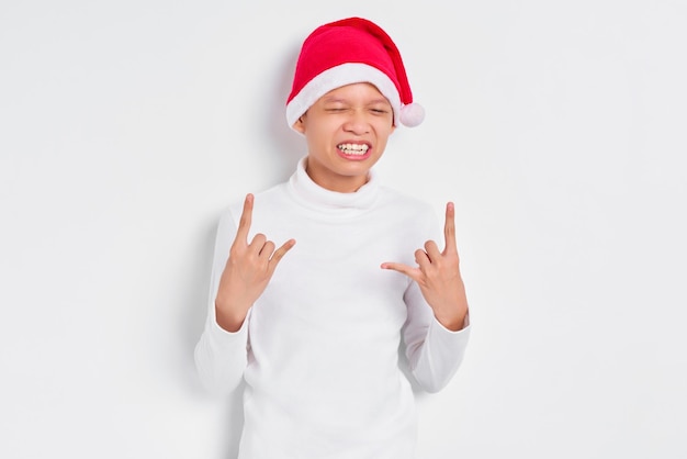 Retrato de homem asiático com chapéu de natal gritando com expressão louca fazendo símbolo de rock com as mãos isoladas em fundo branco