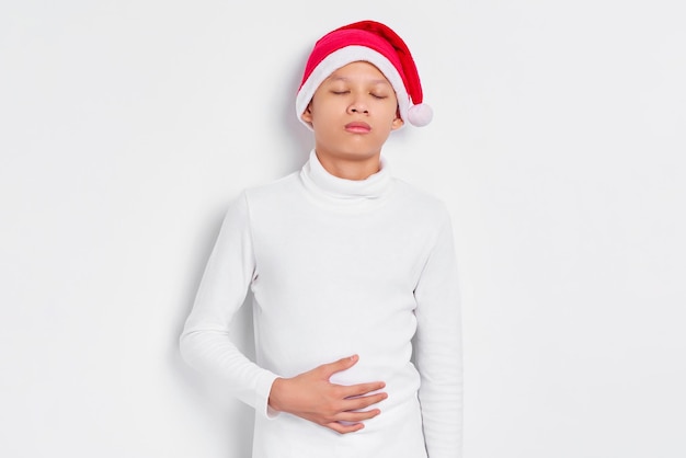 Retrato de homem asiático com chapéu de natal tocando seu estômago sentindo datiety depois de comer uma refeição saudável com bom apetite isolado no fundo branco