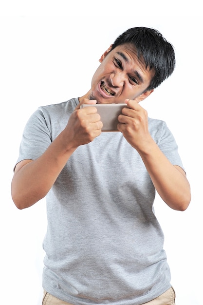 Retrato de homem asiático com raiva vestindo camiseta cinza fica com raiva no smartphone isolado no fundo branco