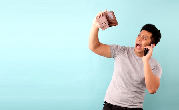 retrato de homem asiático segurando o telefone móvel um chocado, surpreso sem palavras. segurando uma carteira vazia na parede azul.