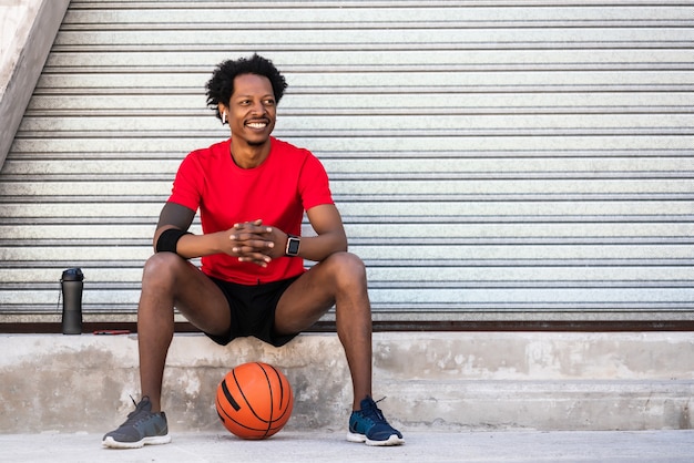 Retrato de homem atleta afro segurando uma bola de basquete e relaxante após o treino enquanto está sentado ao ar livre. esporte e estilo de vida saudável.