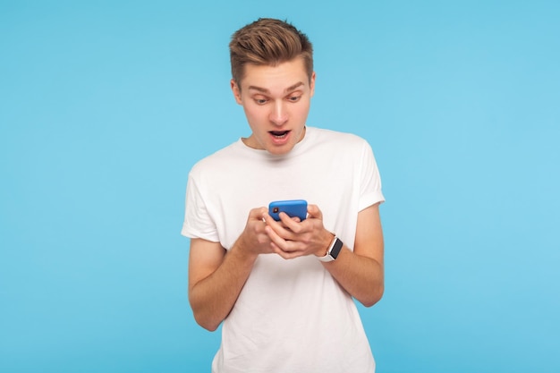 Retrato de homem atônito espantado em camiseta branca usando celular com expressão de surpresa chocado com mensagem na rede social comunicação móvel estúdio interno tiro isolado em fundo azul