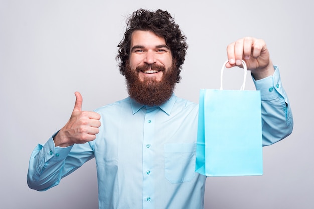 Retrato de homem barbudo alegre em casual mostrando o polegar para cima gesto e sacola de compras azul