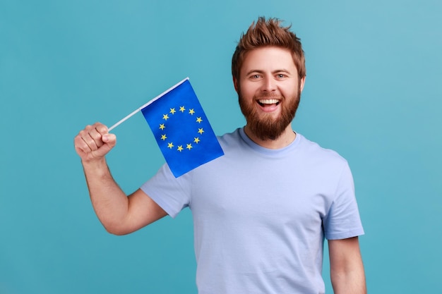 Retrato de homem barbudo animado sorrindo amplamente e segurando a bandeira da união europeia, símbolo da europa, associação da ue e comunidade. tiro de estúdio interior isolado sobre fundo azul.