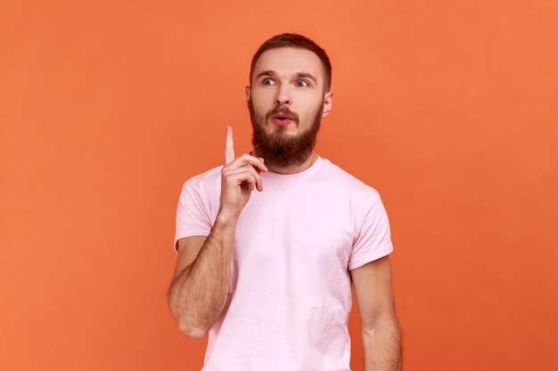 Retrato de homem barbudo bonito inspirado apontando o dedo para cima e parecendo espantado com a ideia genial repentina, obteve solução, vestindo camiseta rosa. Tiro de estúdio interior isolado em fundo laranja.