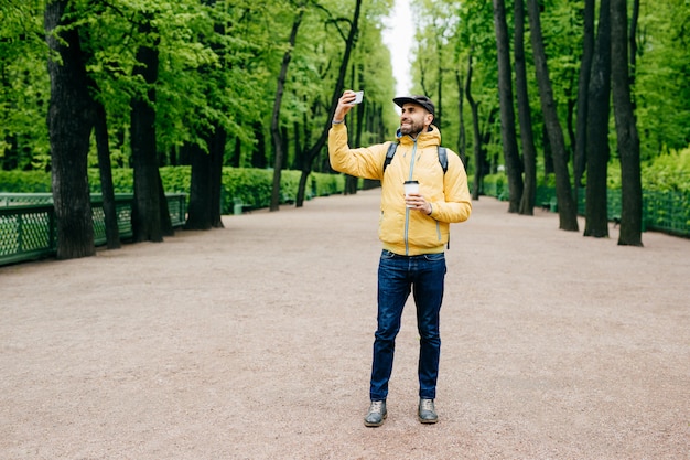 Retrato de homem barbudo bonito no anoraque amarelo, boné e calça jeans tendo expressão feliz enquanto posava no parque fazendo selfie tendo andar bebendo café.